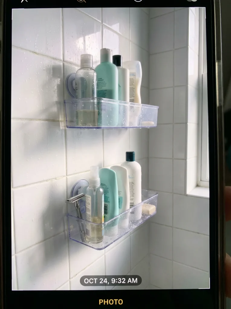 Two clear suction-mounted shelves on a white tile shower wall holding shampoo and conditioner bottles, a spray bottle, a razor, and a bar of soap, with water droplets on the tile and a window letting in light