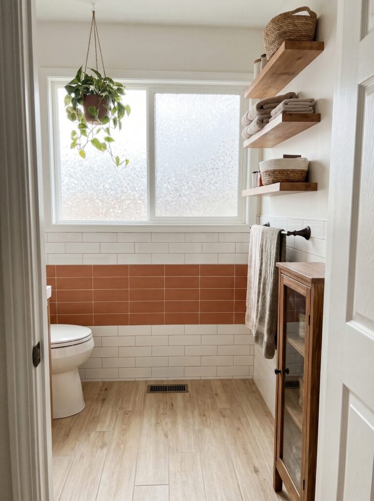 bright bathroom with a horizontal terracotta tile band between white subway tile, wood floating shelves with folded towels, a hanging pothos, and a wood cabinet