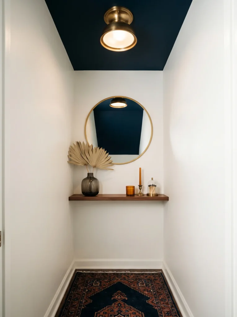 Narrow powder room with white walls and a deep navy painted ceiling, brass flush-mount light, round gold mirror, walnut floating shelf, navy Persian rug