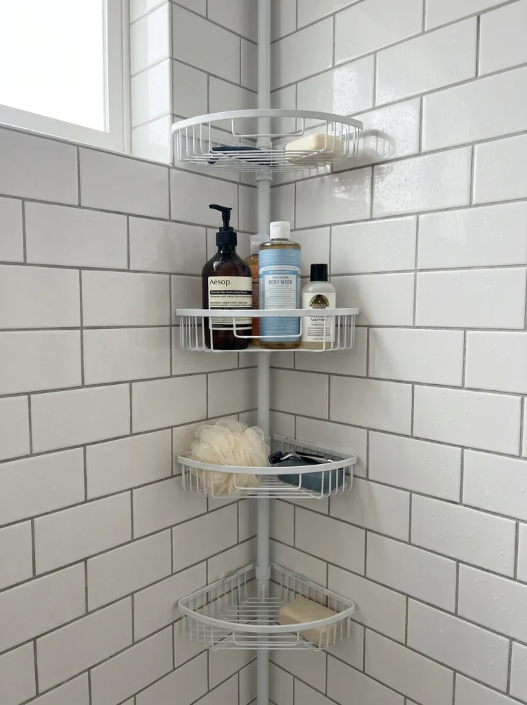 white tension-pole corner shower caddy wedged between two tiled shower walls holding shampoo and body wash bottles at eye level