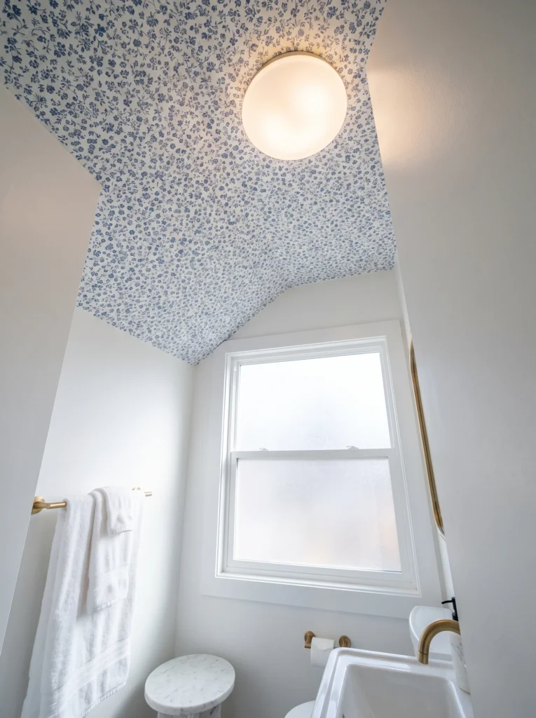 Small blue floral peel and stick wallpaper covering a bathroom ceiling, with a frosted globe light, white walls, frosted window, and gold towel bar