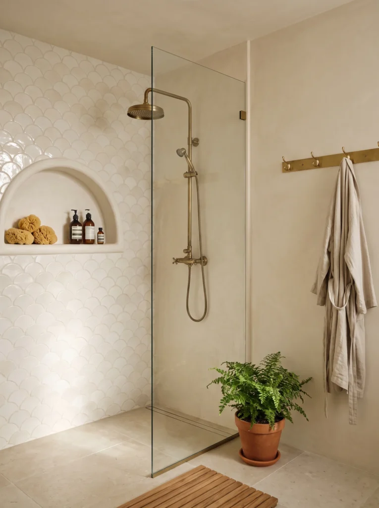 walk in shower with cream fish scale tile, arched niche, brass exposed shower, and teak bath mat
