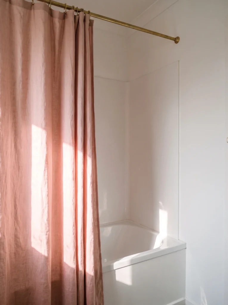 Dusty rose linen shower curtain hanging from a brass rod beside a white bathtub in a bright white bathroom