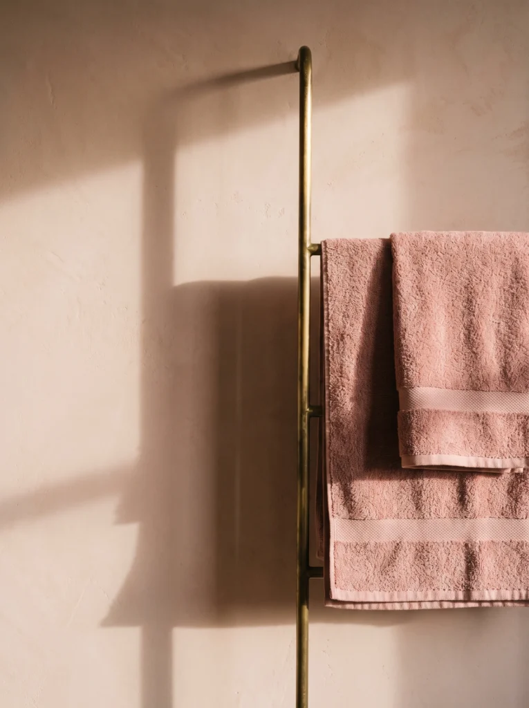 Slim brass towel rail mounted on a pink plaster wall holding folded blush pink towels in afternoon sunlight