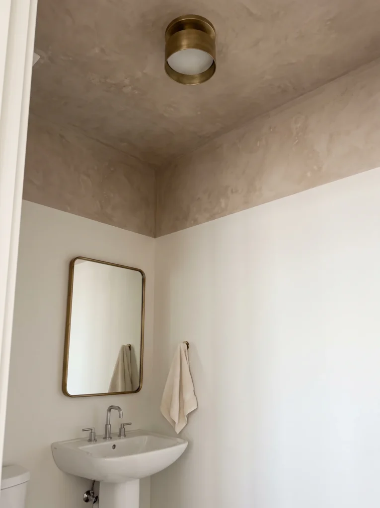 Small bathroom with a mottled taupe limewashed ceiling, brass drum flush mount light, white walls, brass framed mirror and a white pedestal sink