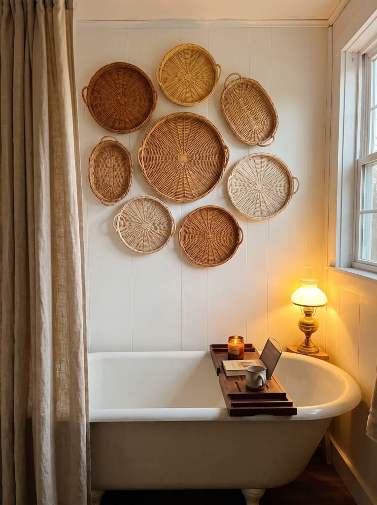 White vertical shiplap wall above a clawfoot tub covered with eight round and oval wicker trays in cream, honey and golden tones, with a wood bath caddy and brass oil lamp