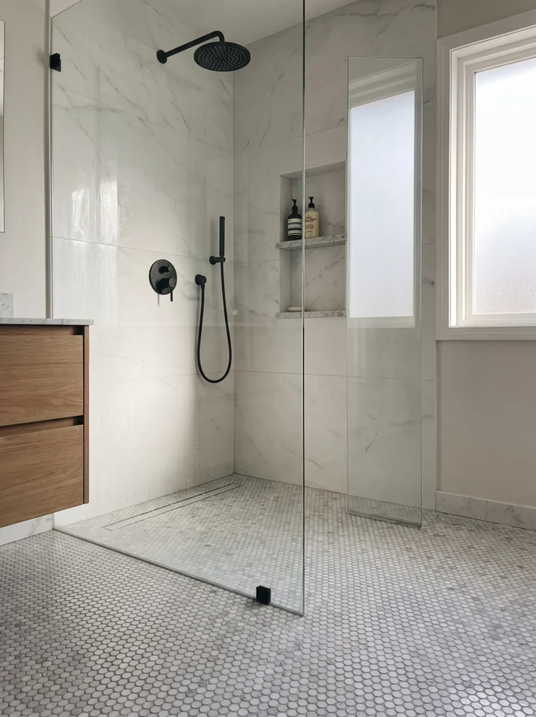 Curbless walk-in shower with marble look wall tile, penny round marble mosaic floor, black fixtures and oak vanity
