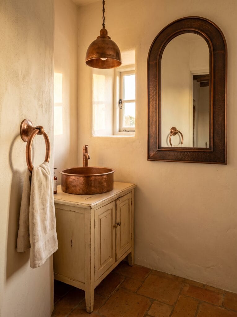 Plaster walled powder room with a hammered copper vessel sink on a distressed cream vanity, arched copper mirror, copper pendant light, copper towel ring and terracotta floor tile