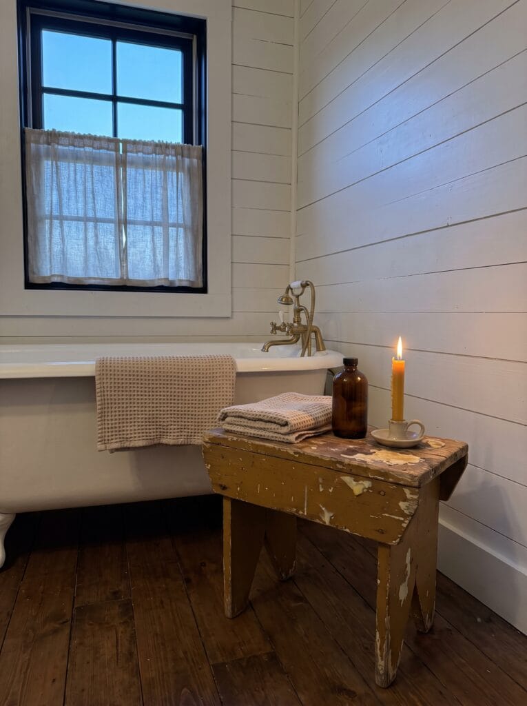 White shiplap bathroom with a black framed window, linen cafe curtain, clawfoot tub with brass telephone faucet and a mustard chippy paint bench holding towels and a lit beeswax candle