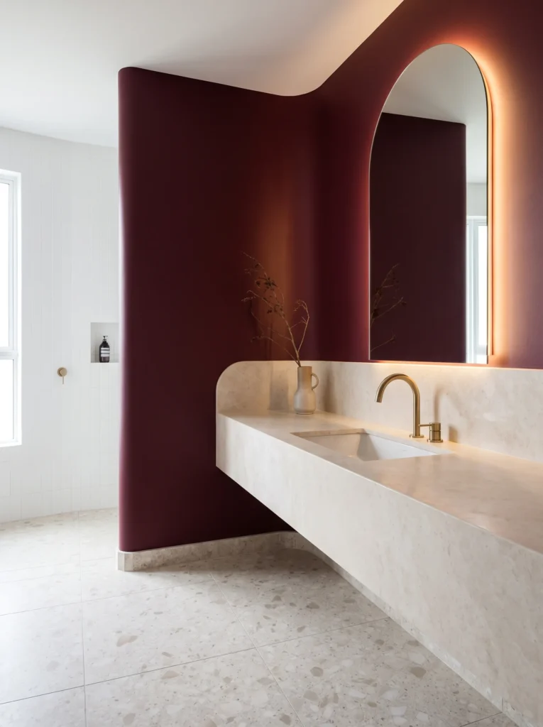 curved burgundy wall with backlit arched mirror, floating stone vanity ledge, brass gooseneck tap, and terrazzo floor