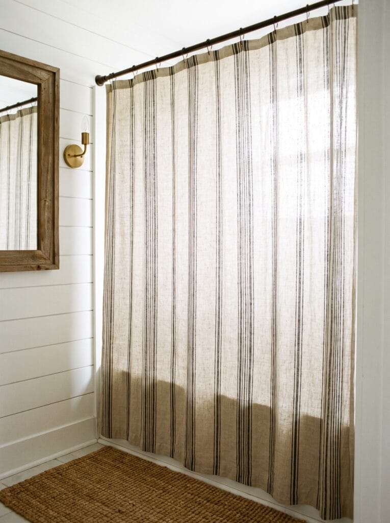Floor to ceiling striped linen shower curtain in cream and black hanging from an iron rod with S hooks, white shiplap walls, rustic wood mirror, brass sconce and a jute bath mat