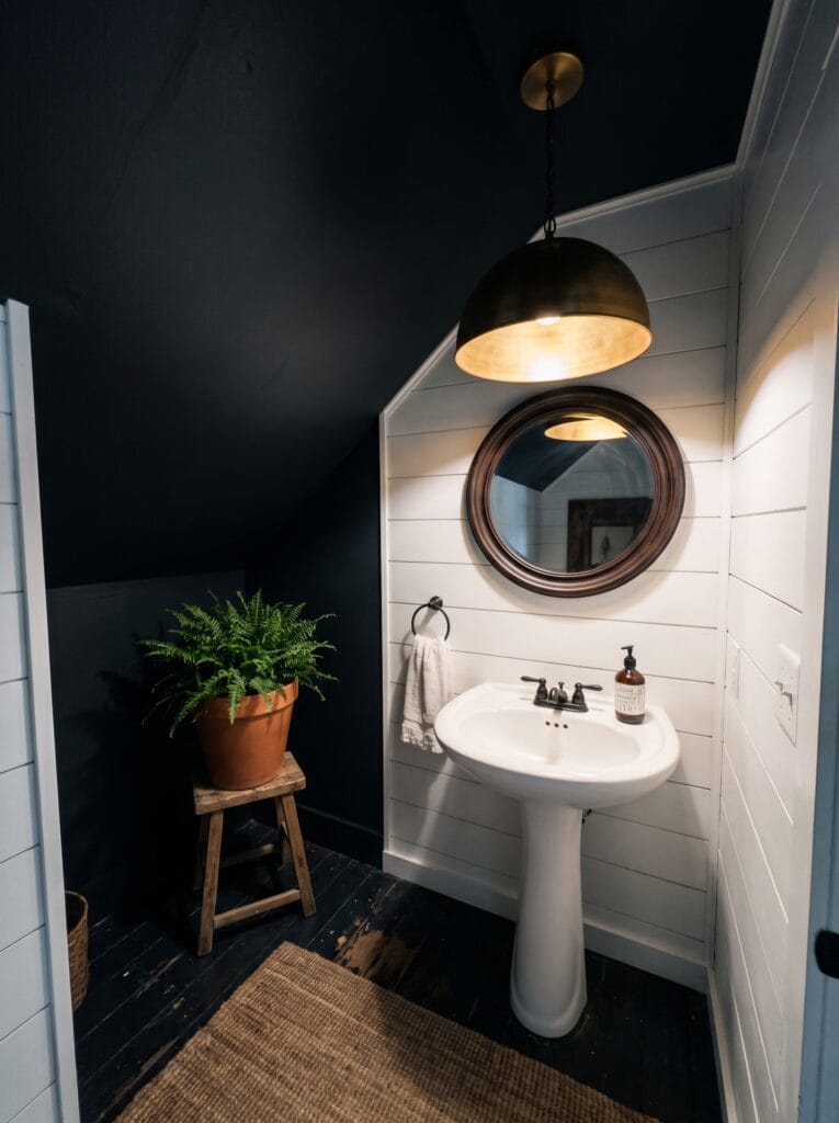 Attic powder room with a dark navy sloped ceiling and walls, white shiplap accent wall, pedestal sink, round wood framed mirror, brass and black dome pendant and a fern on a stool