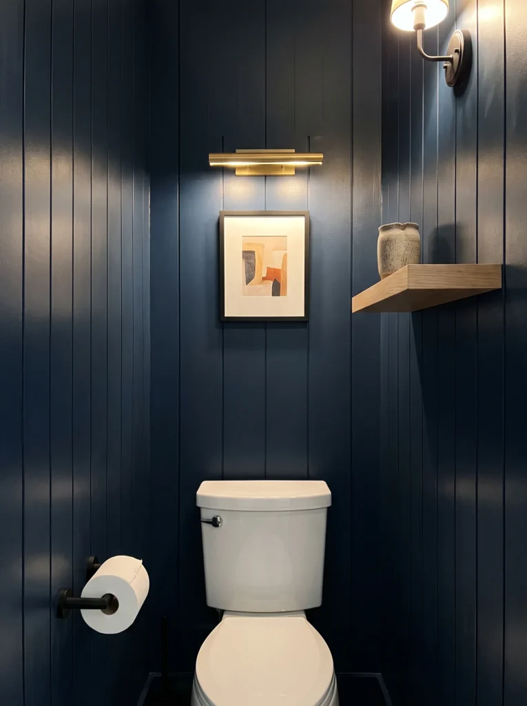 Navy vertical panelled toilet room with framed abstract art, brass picture light and floating oak shelf