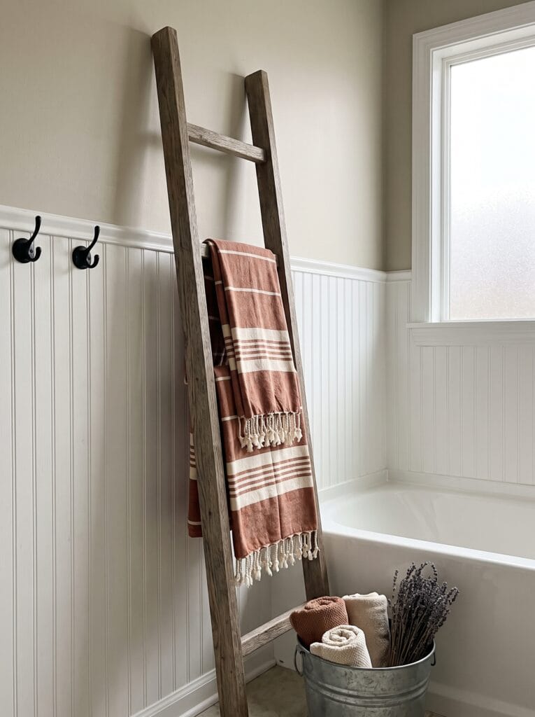 Weathered wood blanket ladder leaning against beadboard wainscoting with rust colored striped Turkish towels, black wall hooks and a galvanized bucket beside a white bathtub