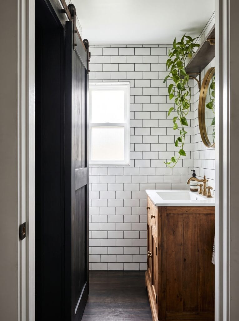 Narrow bathroom with a black sliding barn door on iron hardware, white subway tile with dark grout, dark wood vanity with brass faucet, round gold mirror and trailing pothos