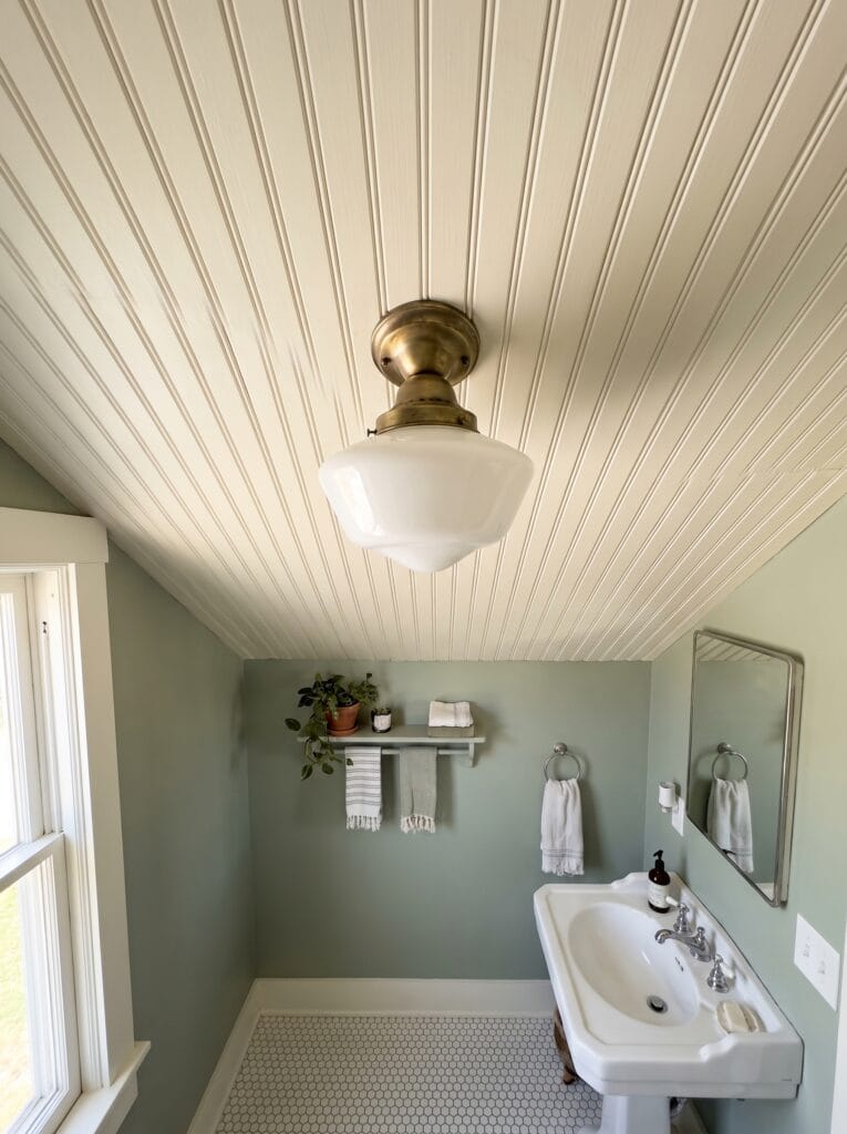 Sage green bathroom under a sloped cream beadboard ceiling with a brass schoolhouse flush mount light, pedestal sink, white penny tile floor and a small towel bar shelf
