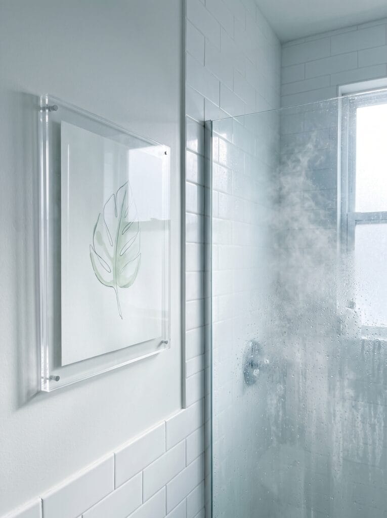 Clear acrylic standoff frame holding a watercolor leaf print beside a steamy glass shower