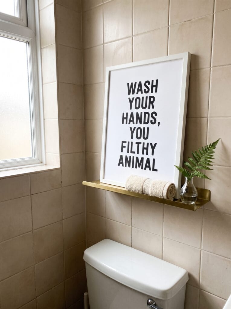 Typography print in a white frame propped on a brass shelf above a toilet