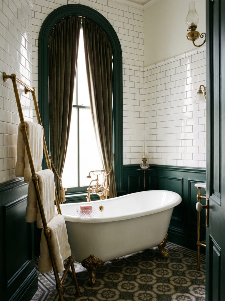 A Victorian bathroom with white subway tile, dark green wainscoting, and a clawfoot tub under an arched window