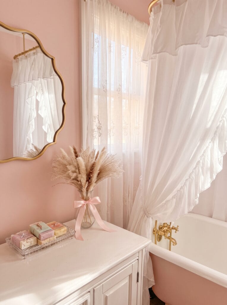 A blush pink coquette powder room with a scalloped gold mirror and a ruffled shower curtain