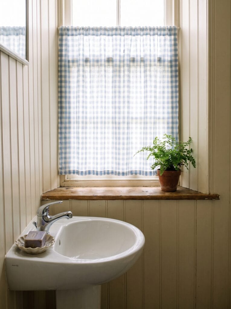 Blue and white gingham cafe curtain at a bathroom window above a pedestal sink