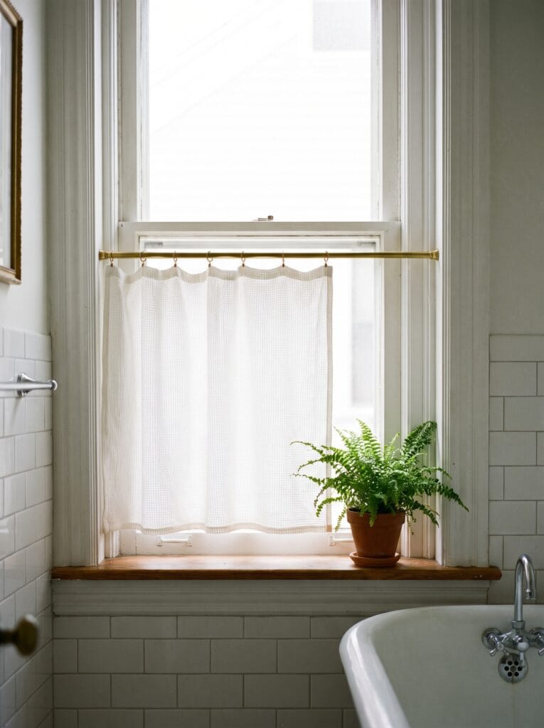 Short white cafe curtain on a brass rod covering the lower half of a bathroom window