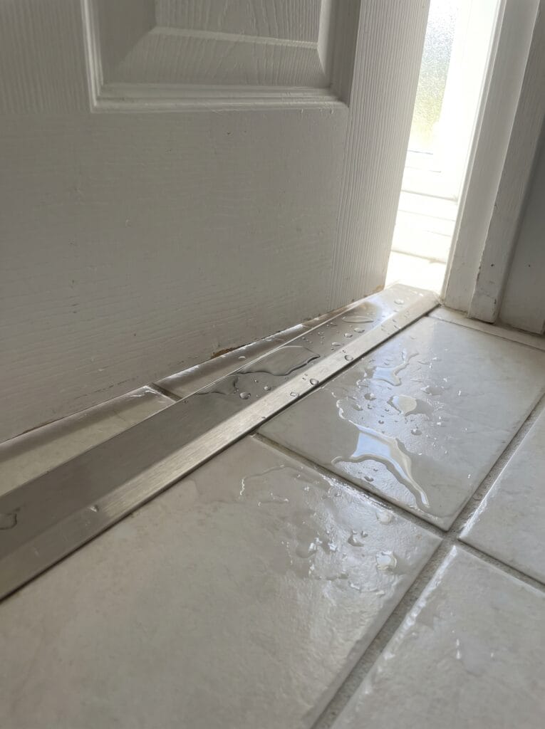 Bottom of a white bathroom door with a brushed metal threshold strip and water on the tile