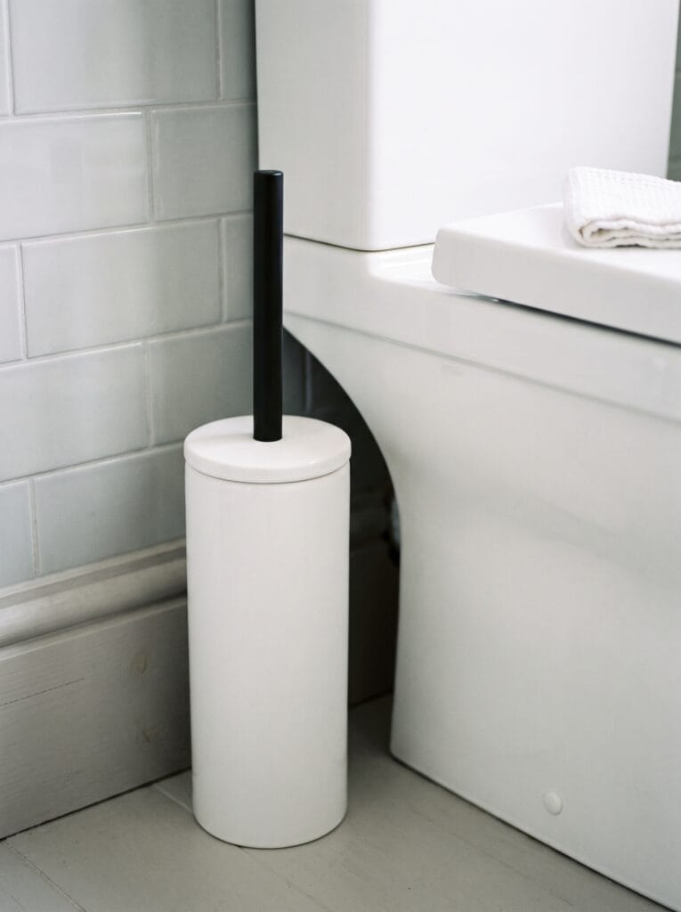 White ceramic toilet brush holder with a fitted lid and matte black handle beside a toilet base