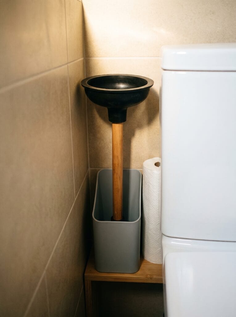 Black rubber flange plunger with a wooden handle standing upright in a slim grey caddy beside a toilet