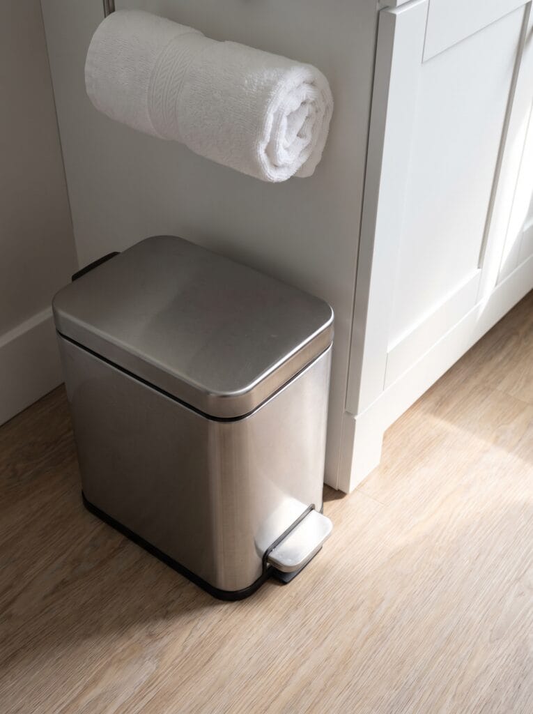 Brushed stainless steel step trash can with a closed lid on wood effect flooring beside a white vanity