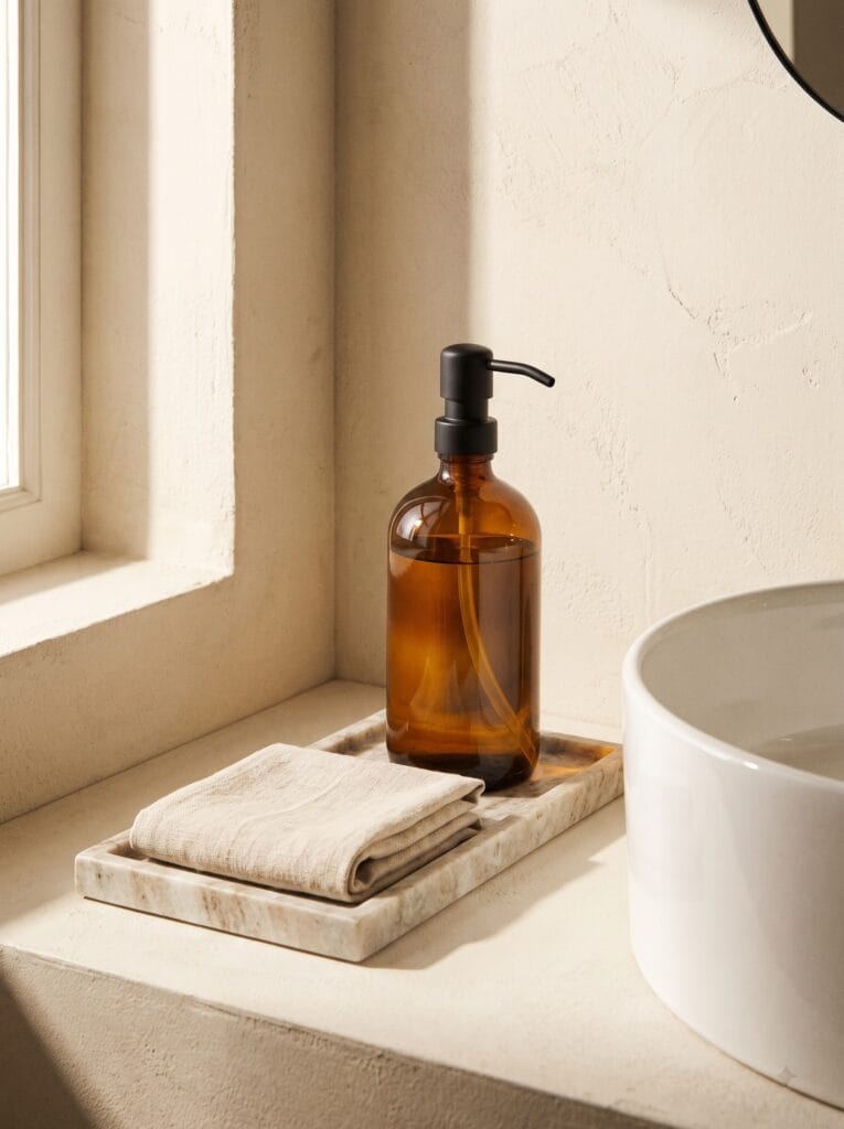 Amber glass soap dispenser with a matte black pump on a marble tray beside a white basin