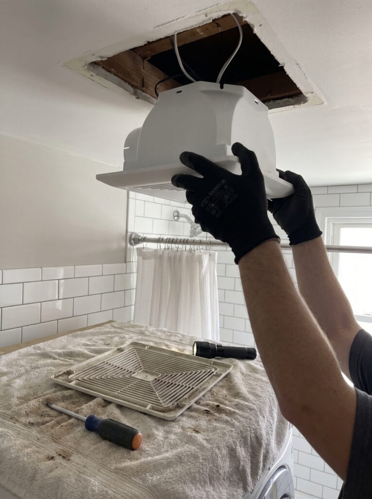 Gloved hands lifting a new white exhaust fan housing into a ceiling opening from inside the bathroom