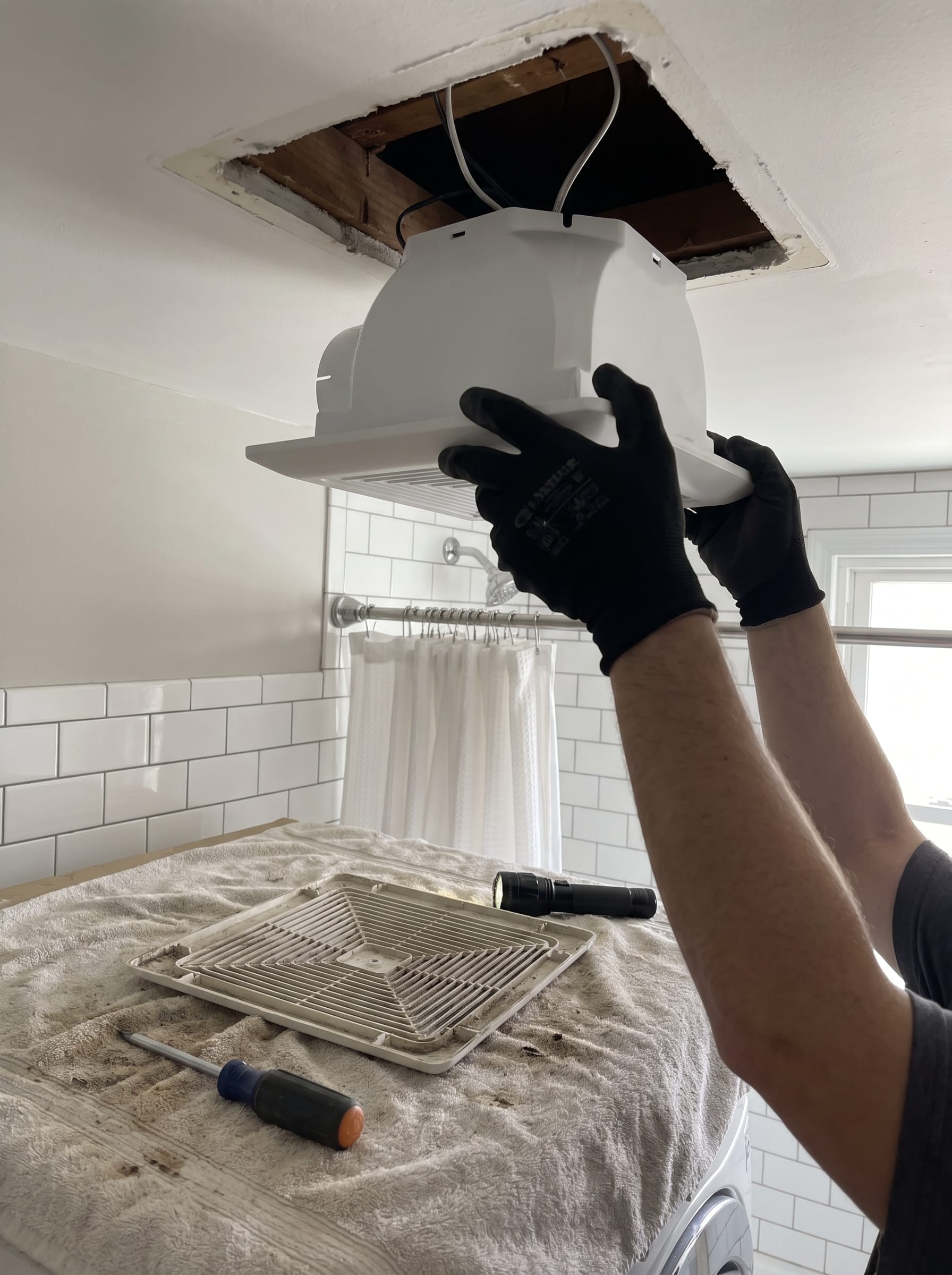 Gloved hands lifting a new white exhaust fan housing into a ceiling opening from inside the bathroom