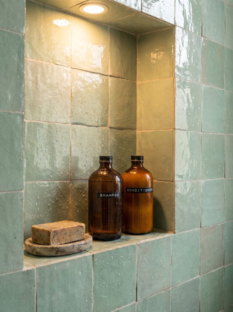 A recessed niche in a walk-in shower tiled in soft green zellige, holding two amber shampoo and conditioner bottles and a bar of soap on a stone dish, with water droplets on the tile and a warm downlight above.