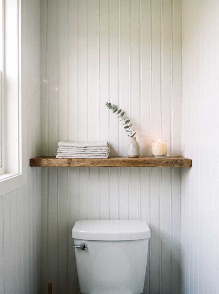 A shallow reclaimed oak shelf mounted above a white toilet on vertical beadboard panelling, holding a stack of folded towels, a small ceramic vase with a eucalyptus stem, and a lit candle, with daylight from a window on the left.