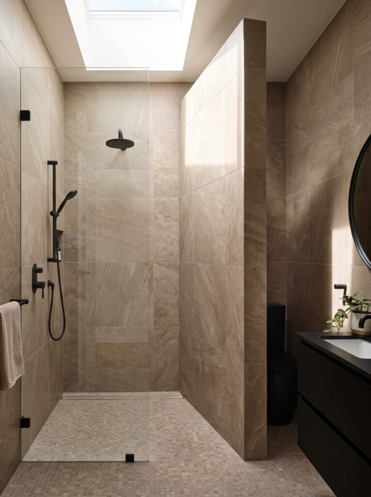 A narrow bathroom with a stone tiled wall screening the toilet, a clear glass shower panel, a skylight overhead and a black vanity with a round mirror.