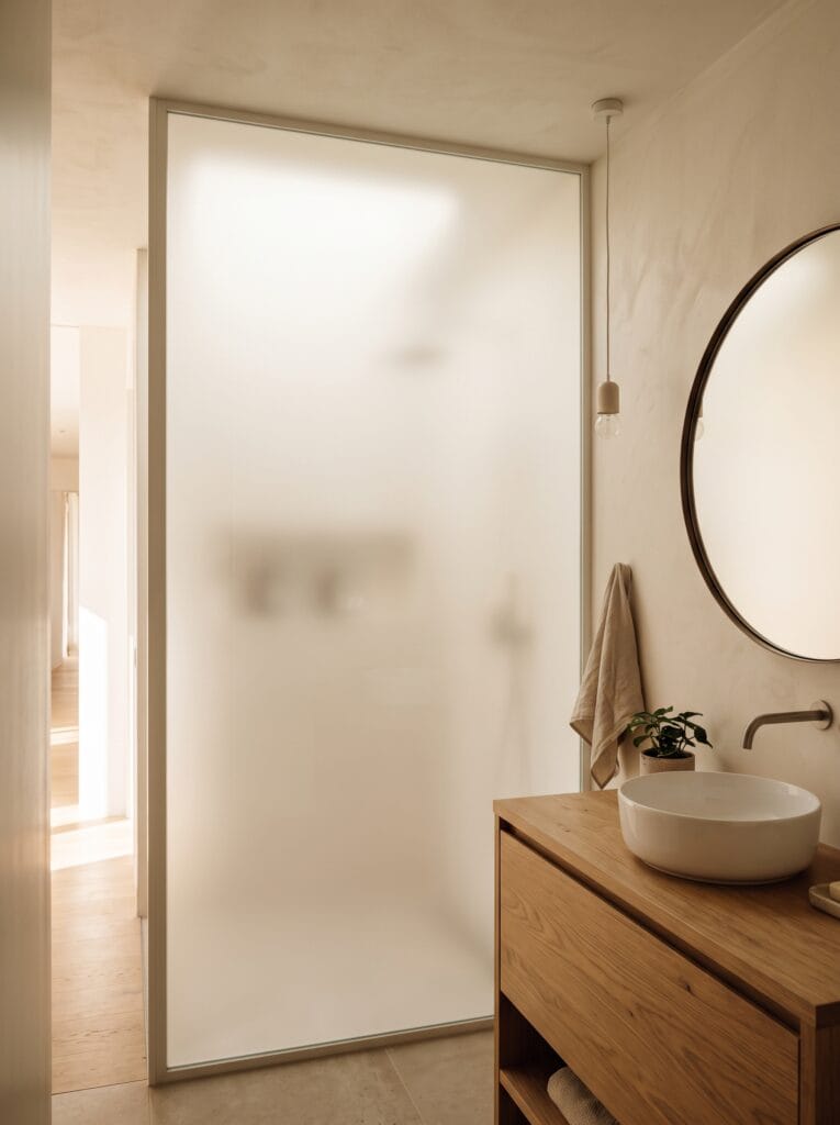 An obscured glass interior partition between a windowless bathroom and a brighter hallway with wood floors, glowing softly beside an oak vanity with a round mirror and a pendant light.
