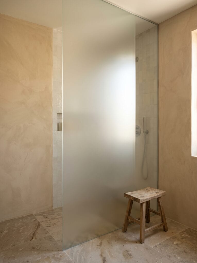A full-height acid-etched glass shower partition glowing with diffused daylight in a warm plaster bathroom with a stone floor and a teak stool.