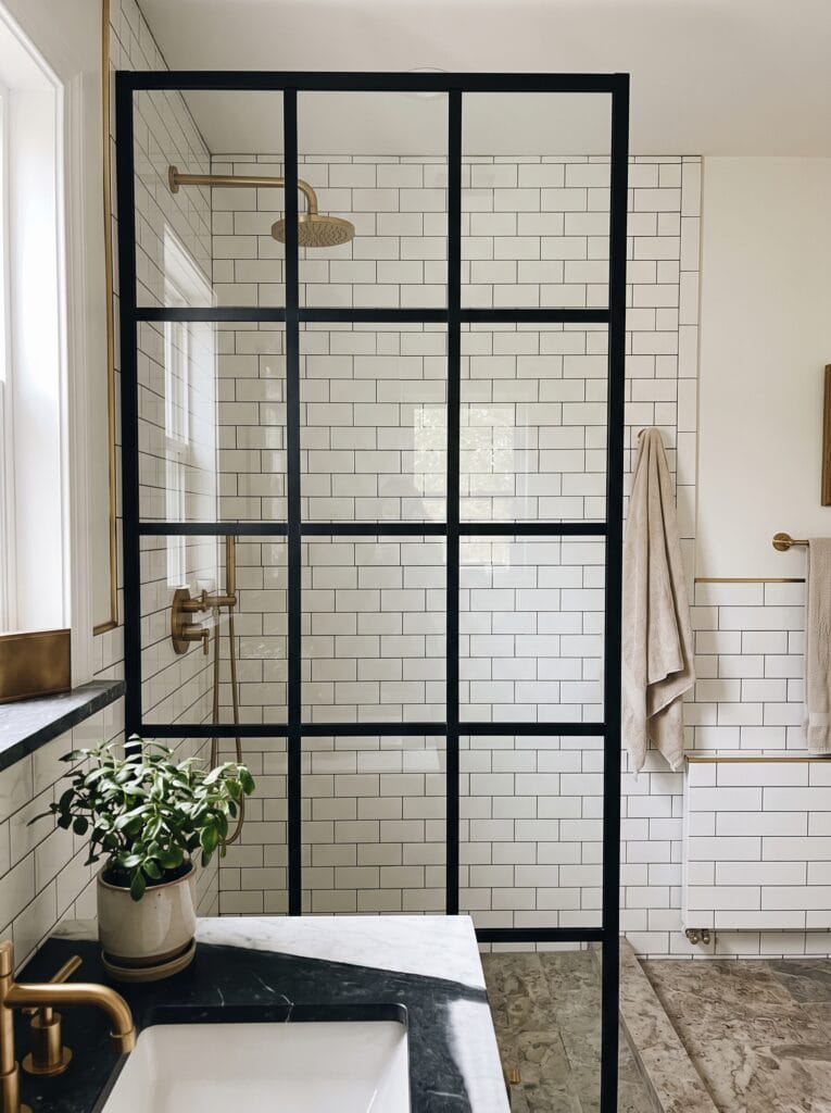 A floor-to-ceiling black-framed grid glass shower partition against white subway tile with brass fixtures, a plant on a dark stone vanity in the foreground.