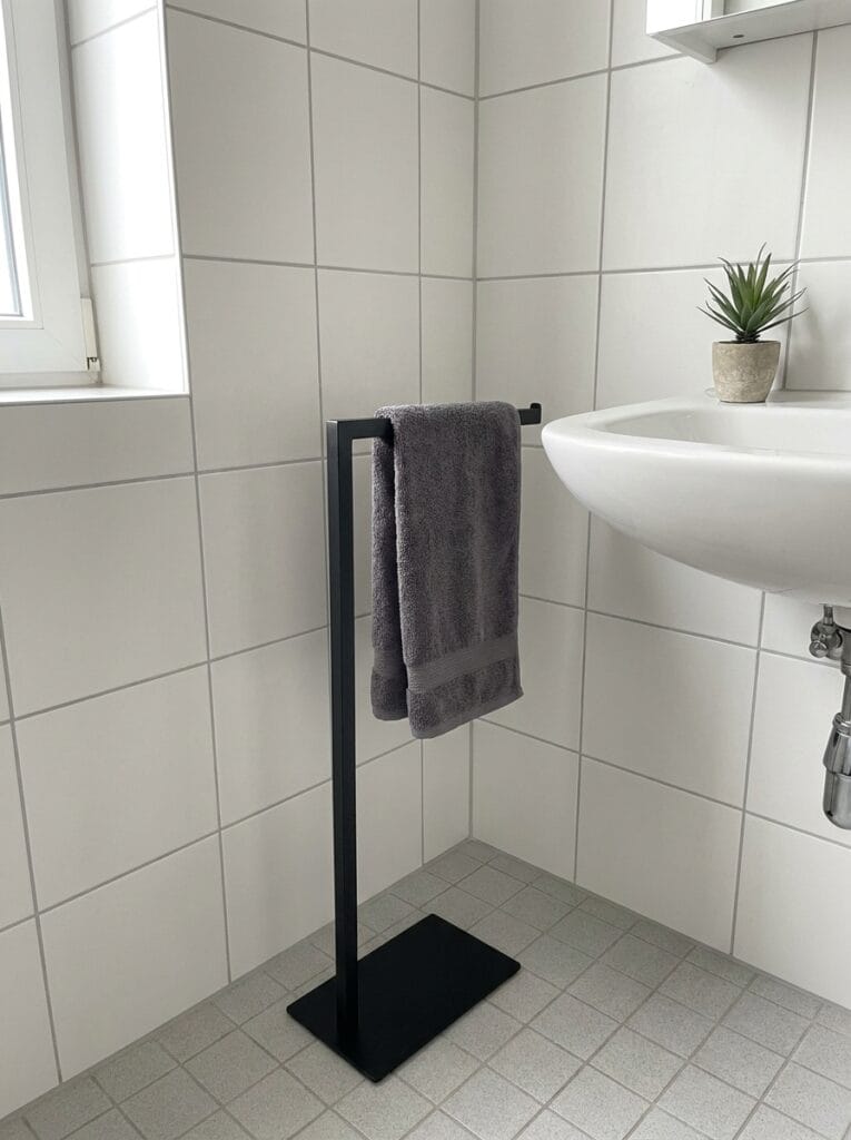Slim matte black freestanding towel stand beside a wall-hung sink in a white tiled bathroom