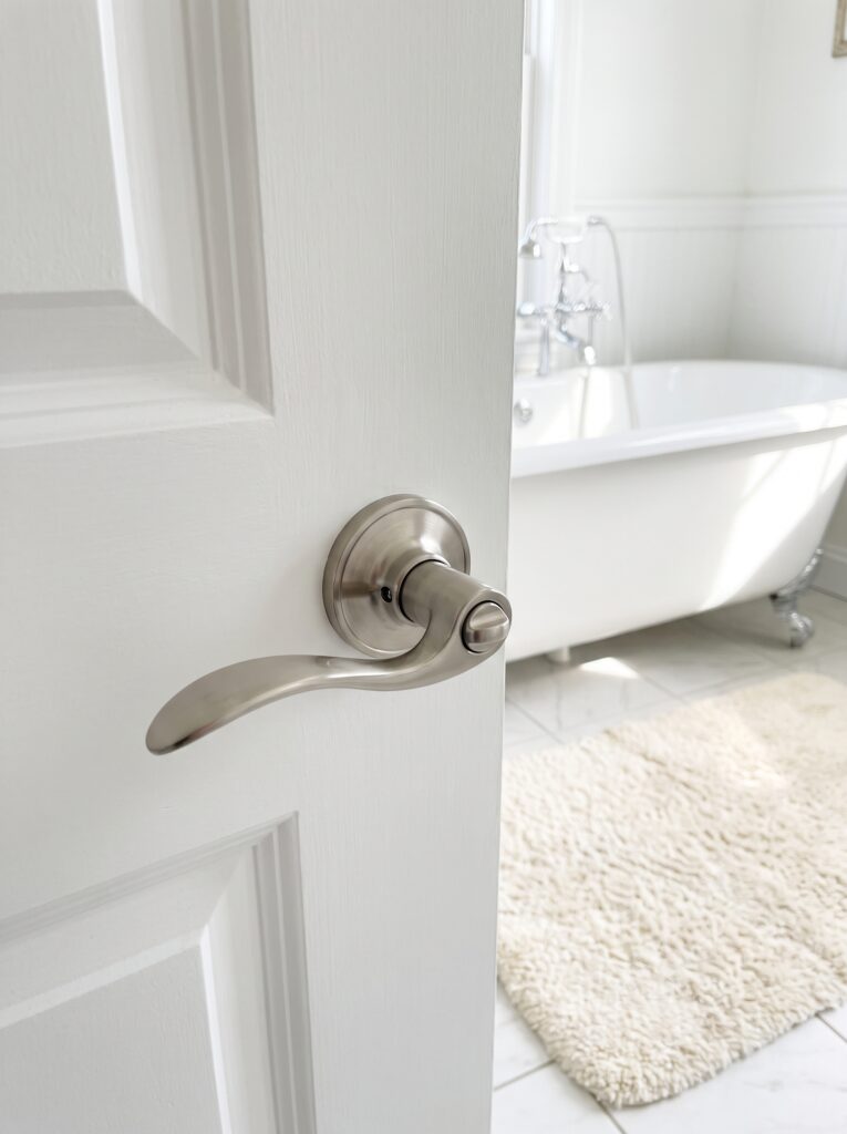Brushed nickel privacy lever on a white panelled bathroom door opened onto a clawfoot tub and cream rug