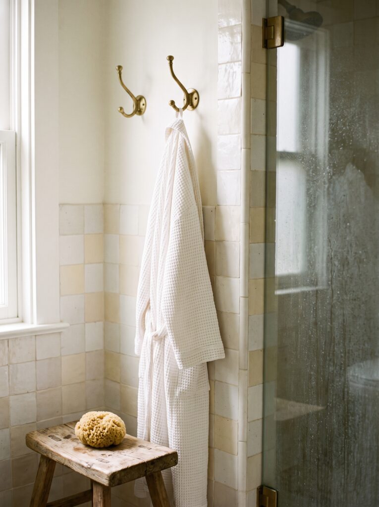 Two antique brass robe hooks on a cream zellige tile wall with a white waffle robe hanging beside a glass shower door