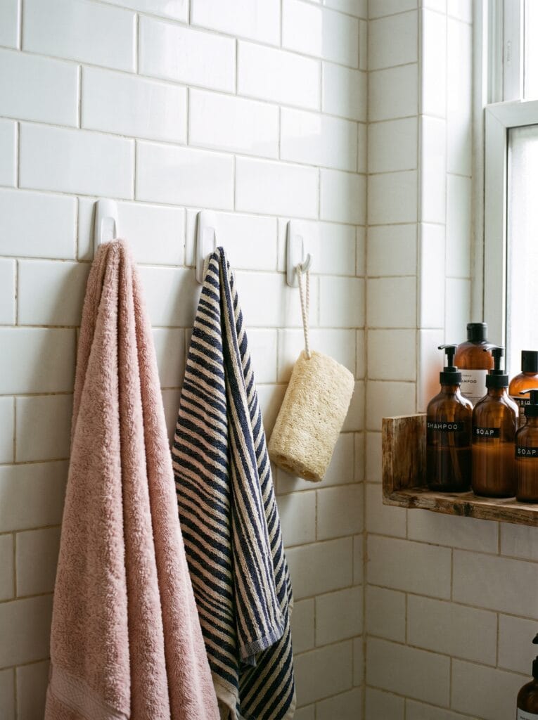 white adhesive hooks on subway tile holding a blush towel, a striped towel and a loofah