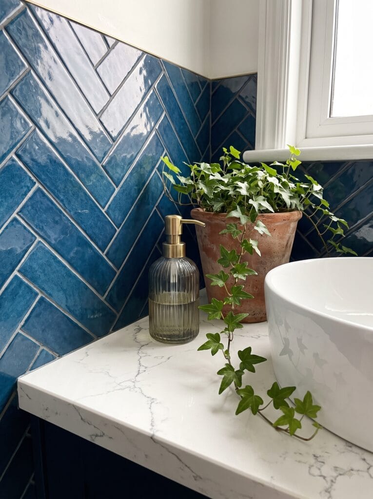A blue herringbone tile backsplash behind a white marble counter with a vessel basin and a potted ivy