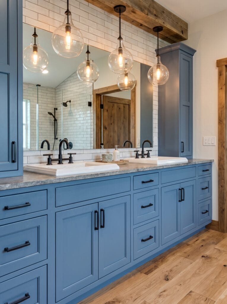 A wide blue double vanity with two white sinks, concrete counter, glass pendant lights and a reclaimed wood beam above