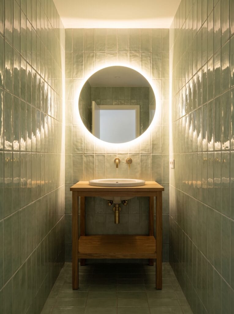 Sage green vertical tile around a backlit round mirror in a windowless bathroom