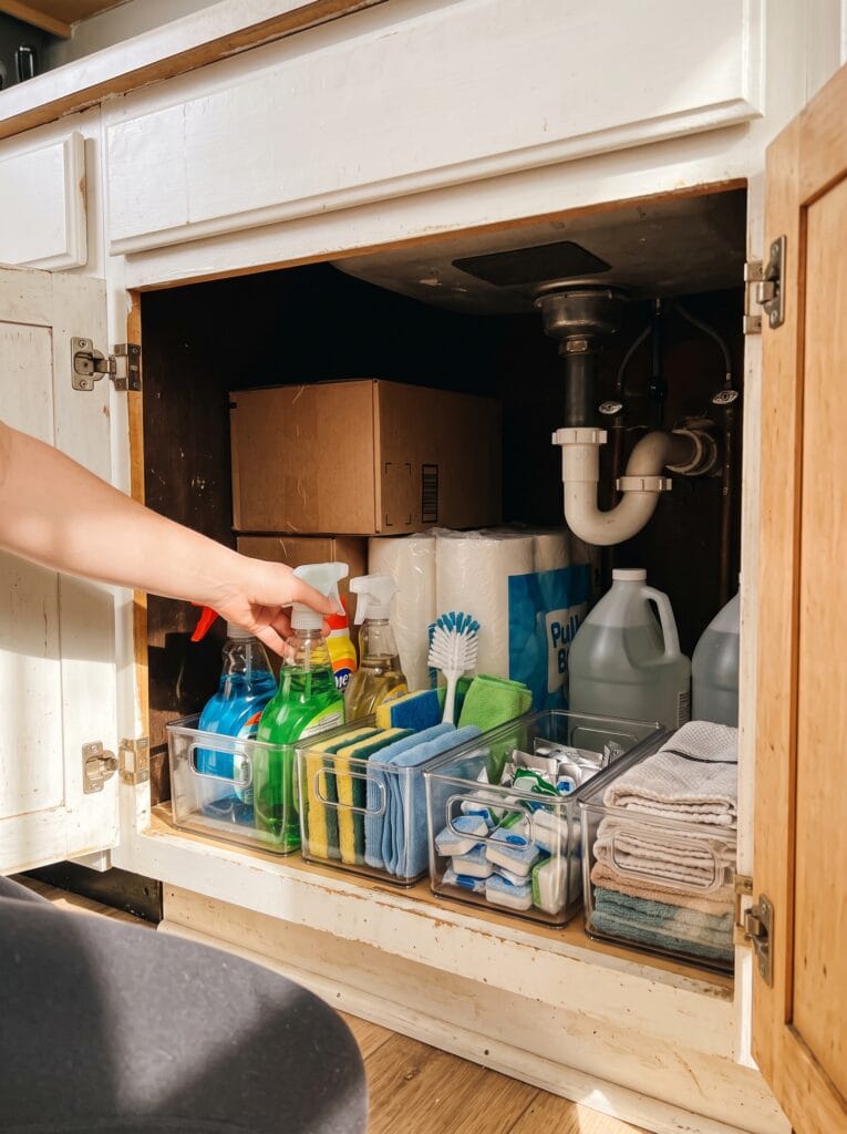 Open under sink cabinet with clear bins at the front and sealed backup boxes pushed to the back