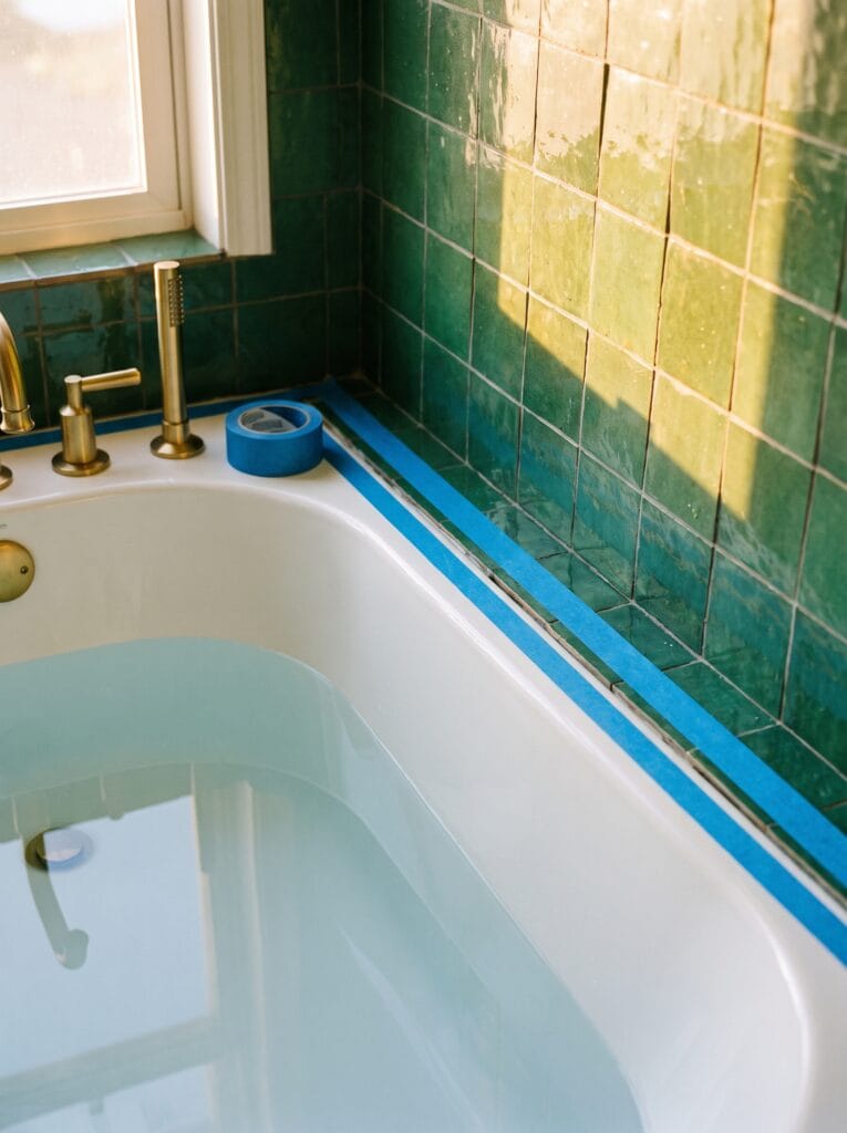 A white bathtub filled with water, with two strips of blue painter's tape along the joint where it meets green zellige tile.