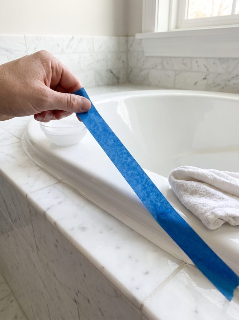 A hand peeling blue painter's tape away from a freshly smoothed white caulk line on a marble tub surround.