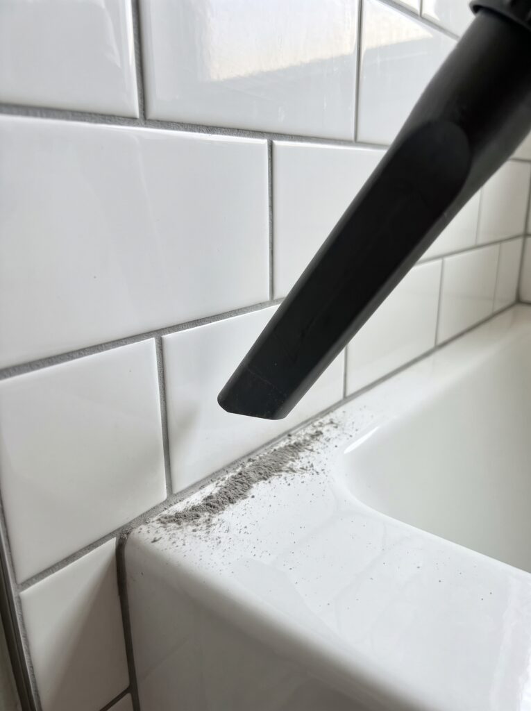 Slim vacuum nozzle above a white tile wall with freshly emptied grout lines and fine dust on the tub ledge